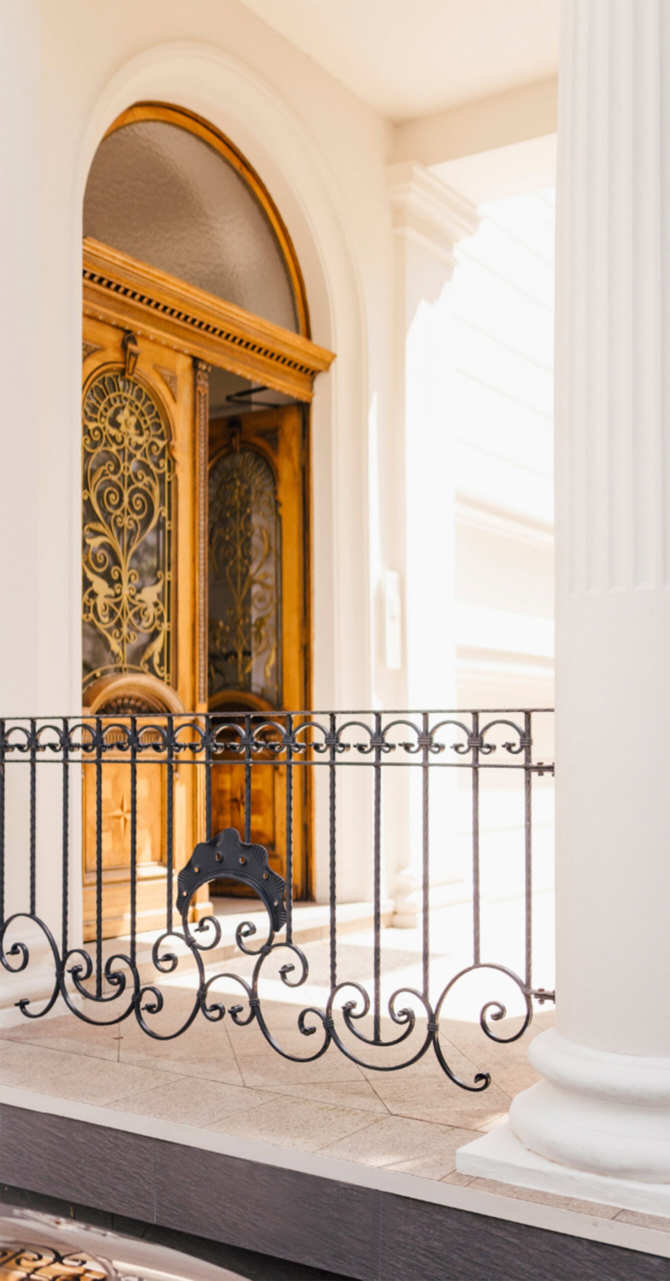 Ornamentale Holztür mit Goldverzierungen hinter schmiedeeisernem Geländer und weißen Säulen.