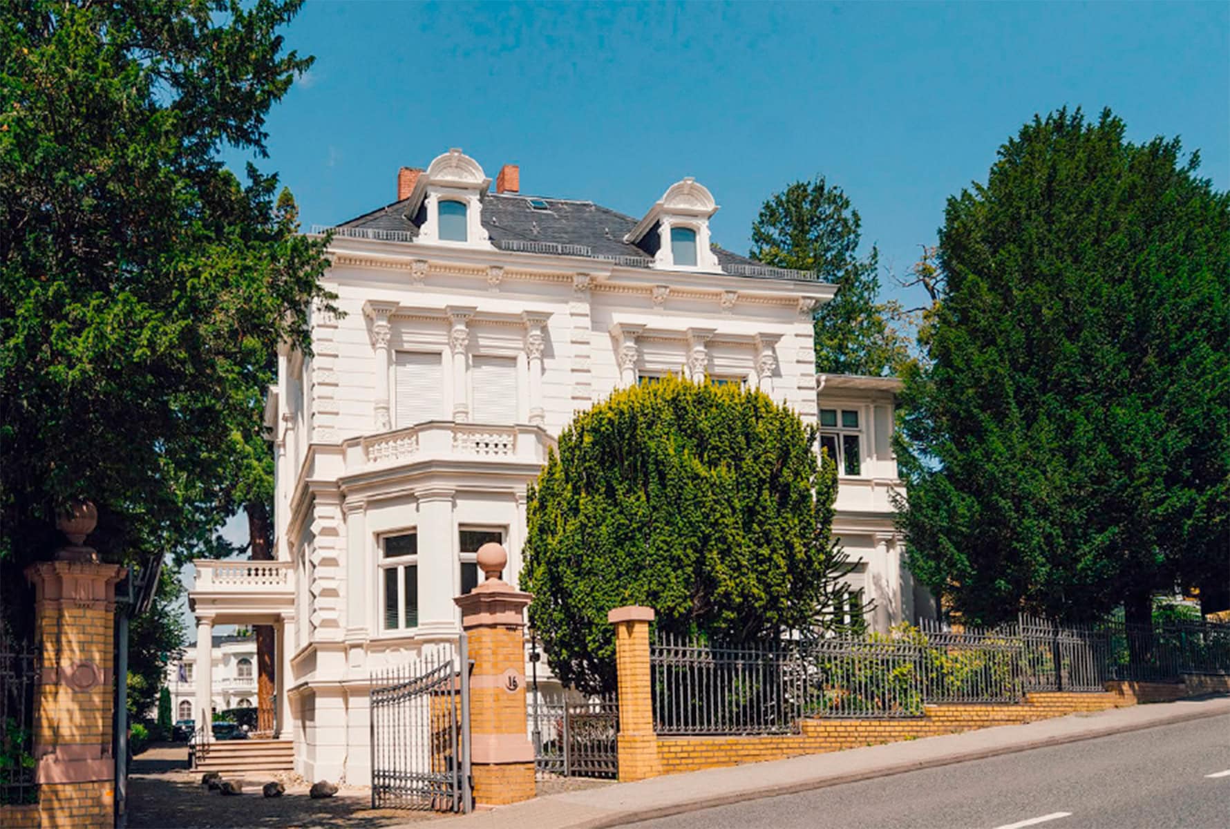 Weiße Gründerzeit-Villa mit Stuckfassade, Gauben und schmiedeeisernem Zaun vor grünen Bäumen.