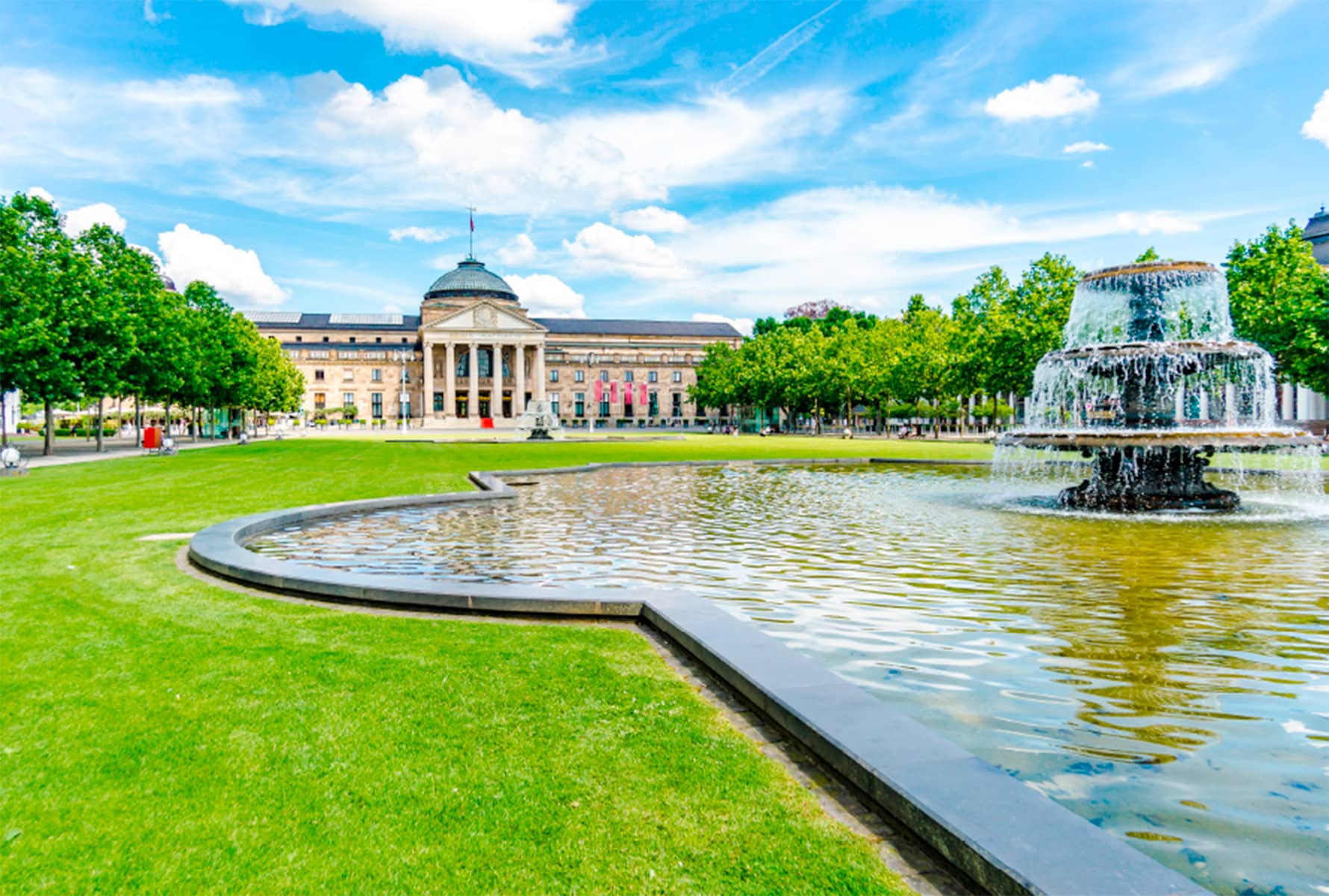 Klassizistisches Gebäude mit Kuppel vor grüner Wiese, Springbrunnen und blauem Himmel mit Wolken.