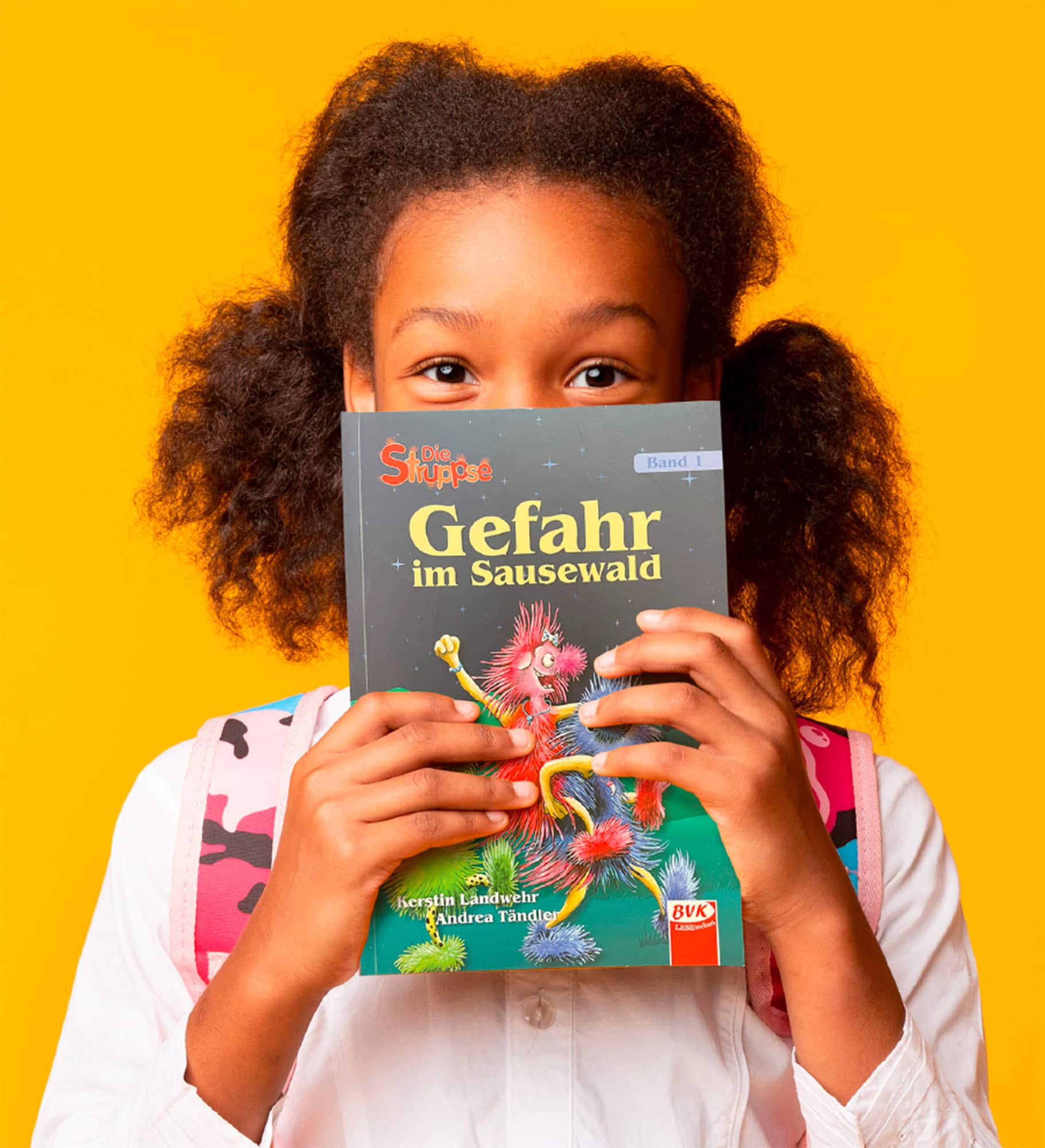 Mädchen mit Zöpfen hält Kinderbuch 'Die Snuppsé – Gefahr im Sausewald' vor dem Gesicht, gelber Hintergrund.